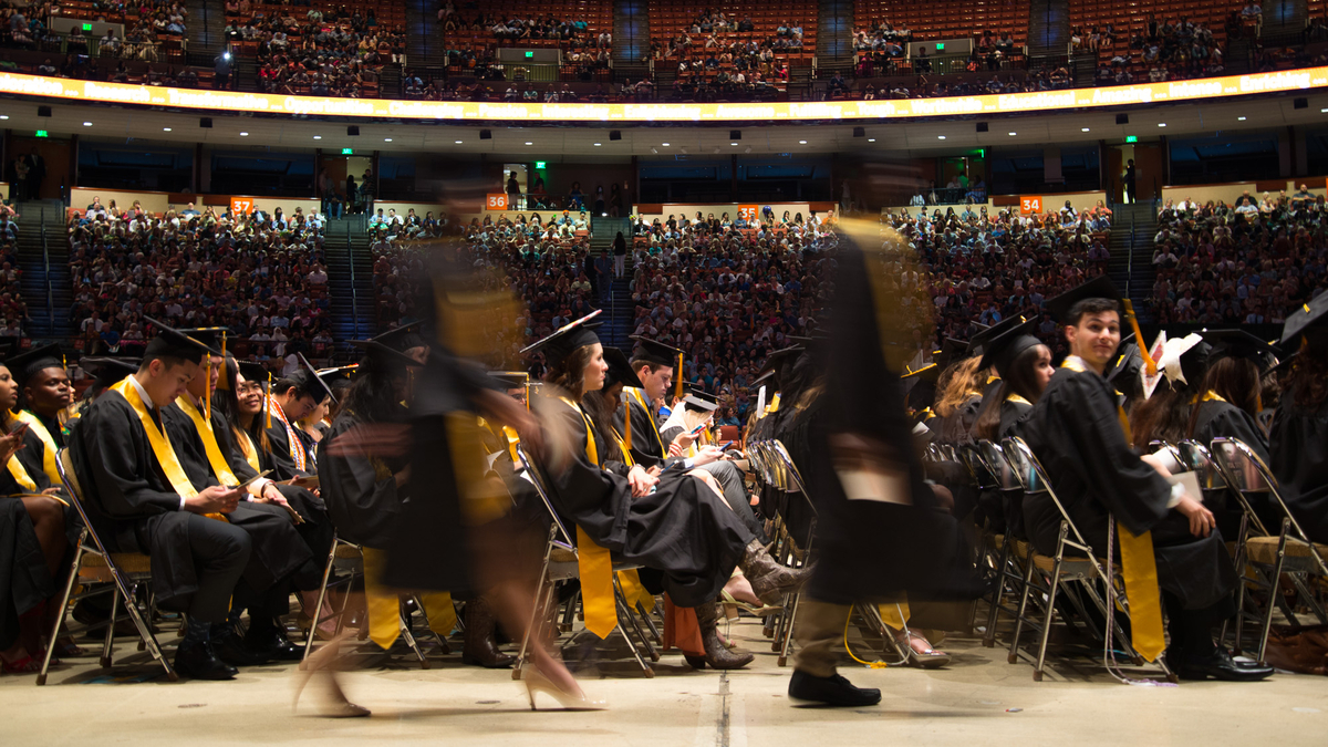 A blur of graduates proceed in a line while other graduates sitting in rows look on in a crowded arena.