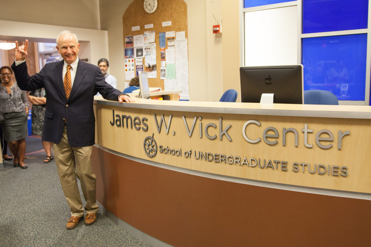 A man in a suit flashes the hook 'em horns hand sign next to a desk where the sign reads "James W. Vick Center, School of Undergraduate Studies"