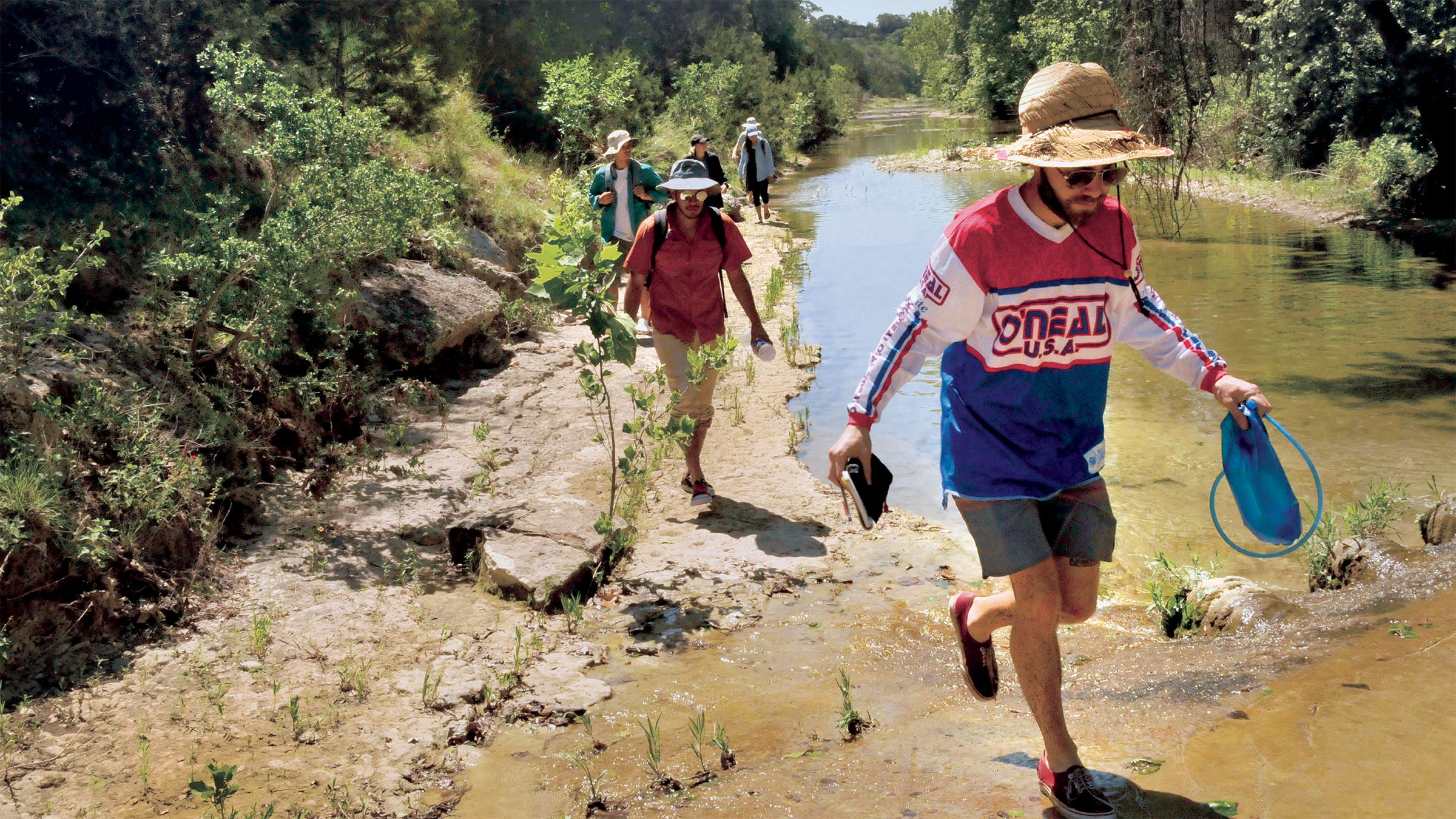 A young man in a hat and sunglasses leads a group of students down a creek with equipment in hand at a field staton