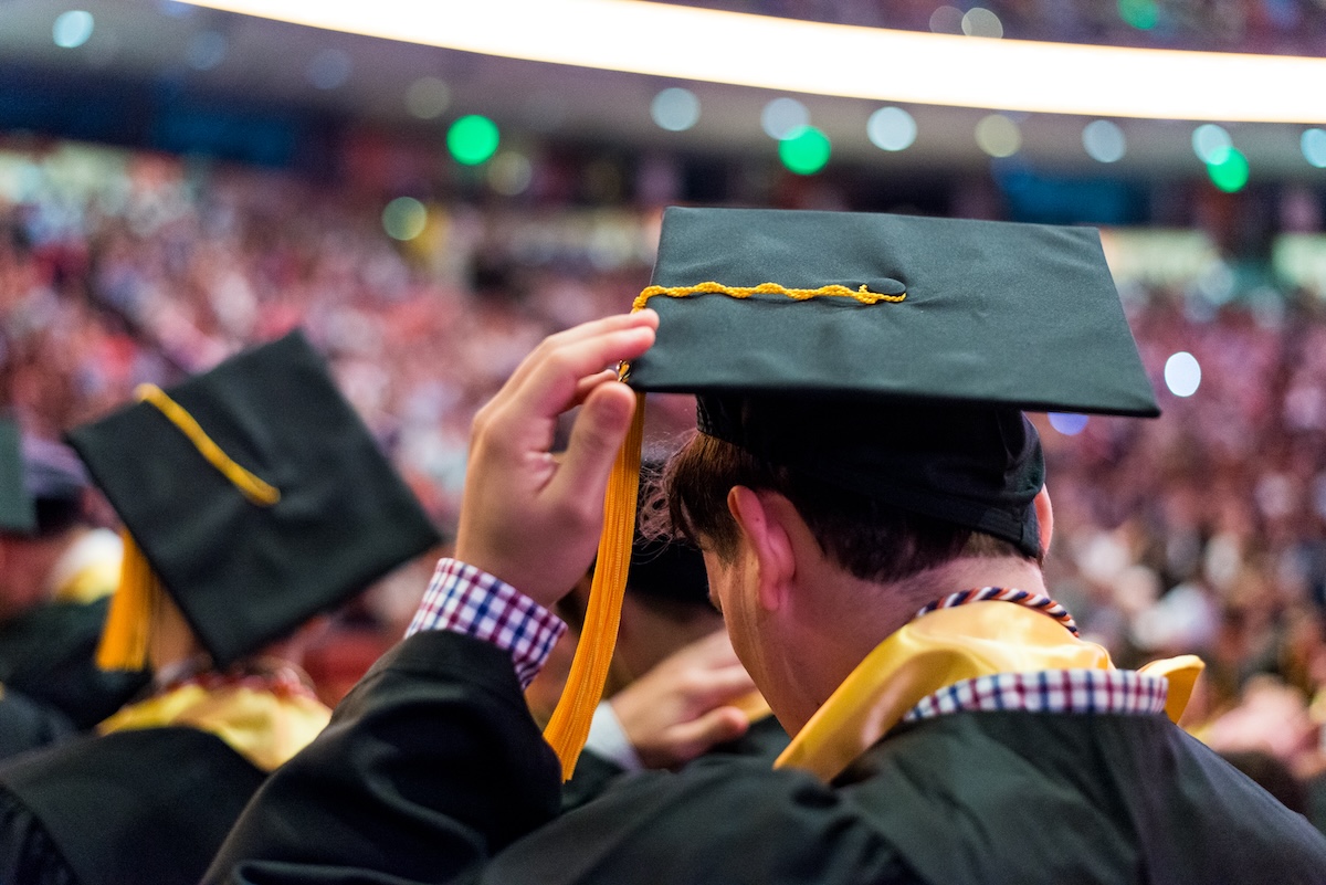 A graduate taps his cap while processing with others in a crowded arena.