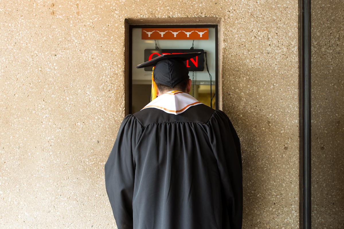 A person in graduation regalia is shown from behind entering an area that displays Longhorn silhouettes and the word open