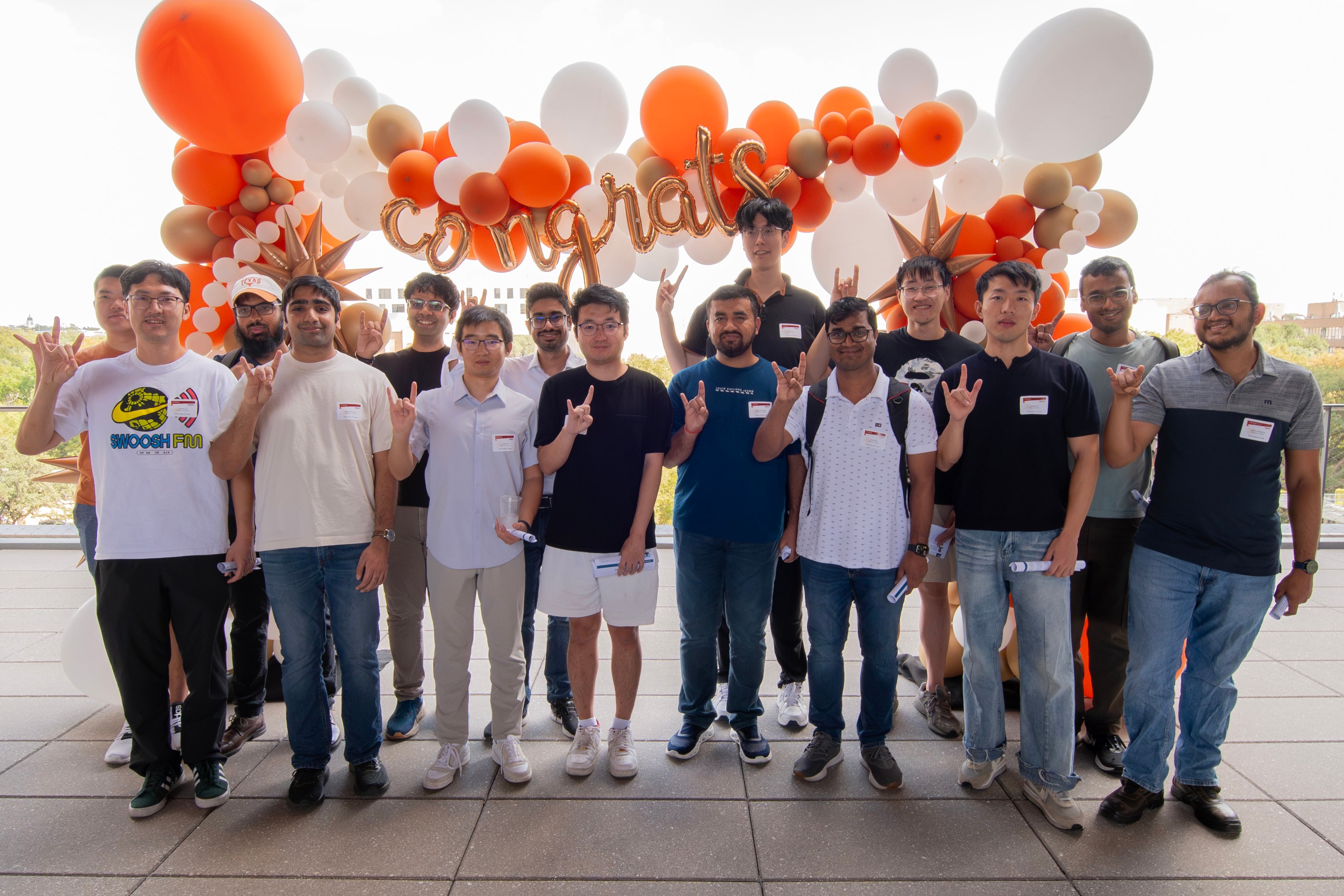 A group of graduate students stand in front of a congratulations message and balloon display, while showing the hook 'em hand signal