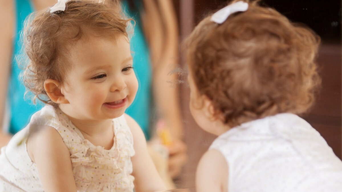 A toddler gazes at herself in the mirror and smiles.