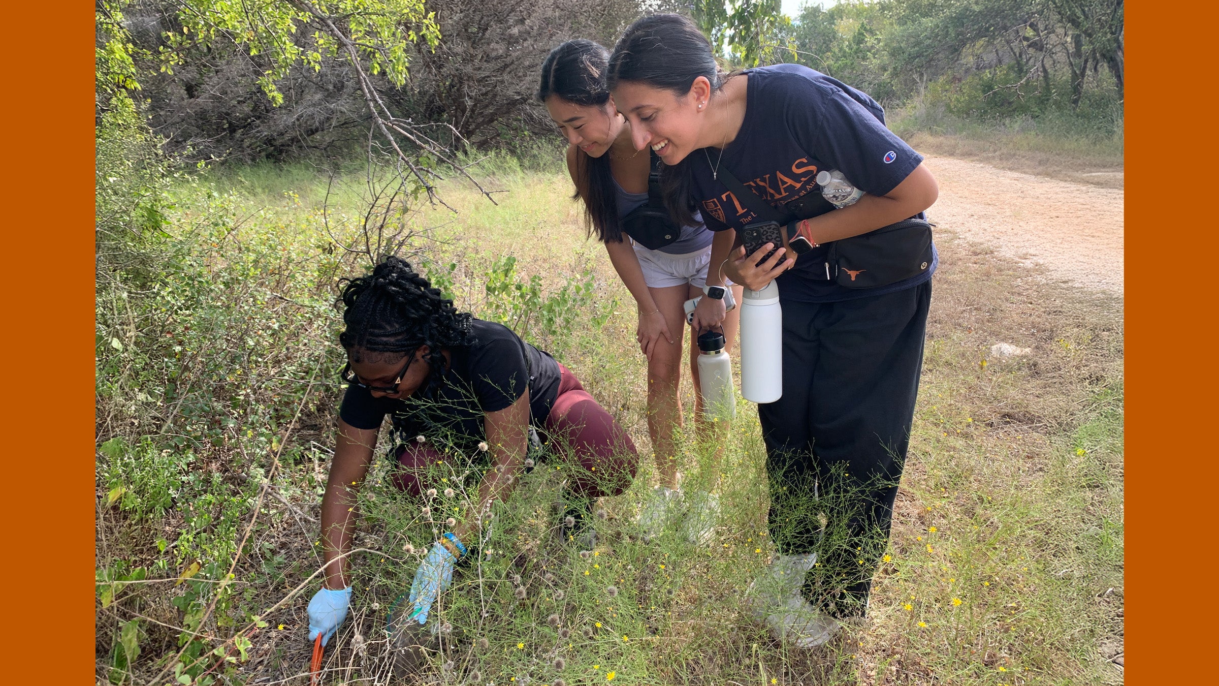 Two young women look on as a third digs in the soil with gloves in an area where wildflowers grow near a path.