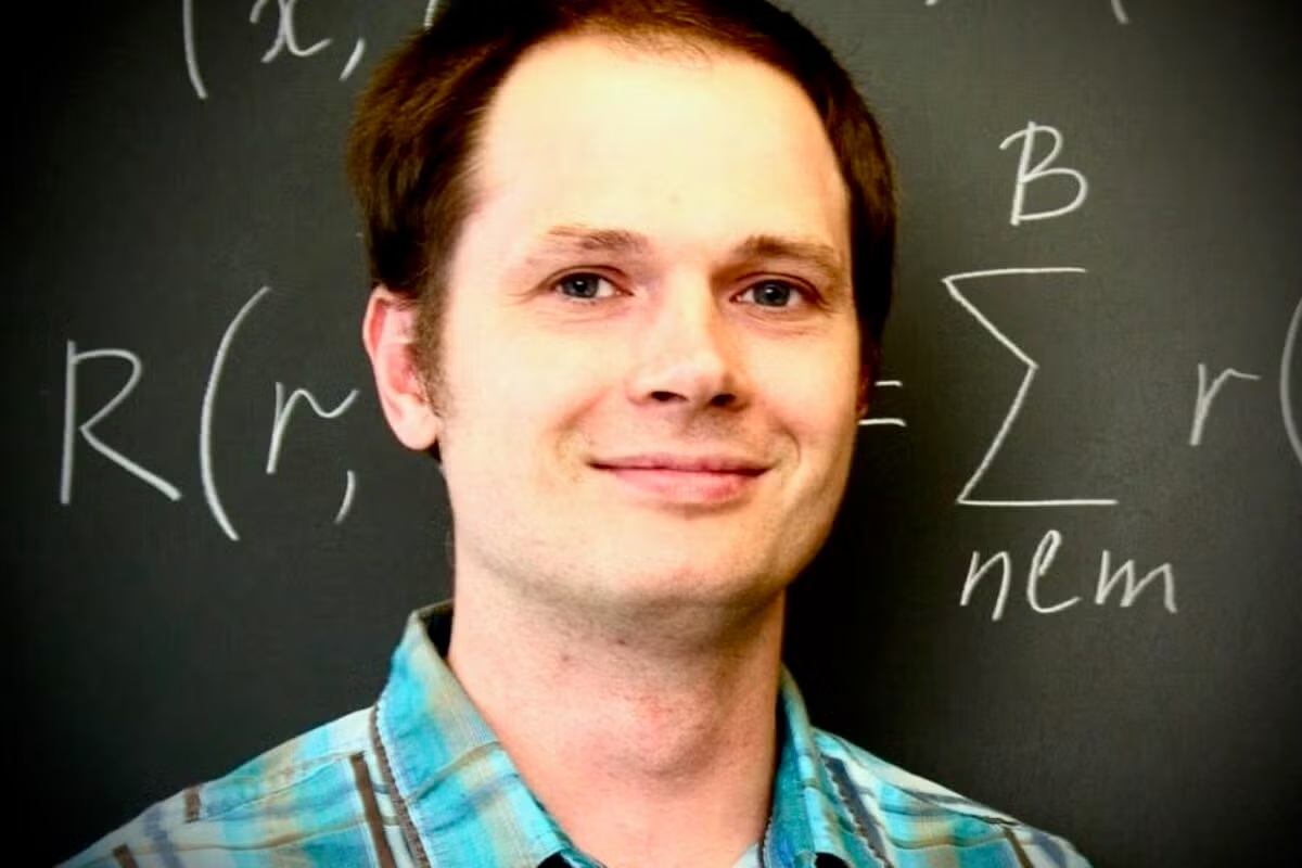 A man smiles, in front of a chalk board with formulas written on it.