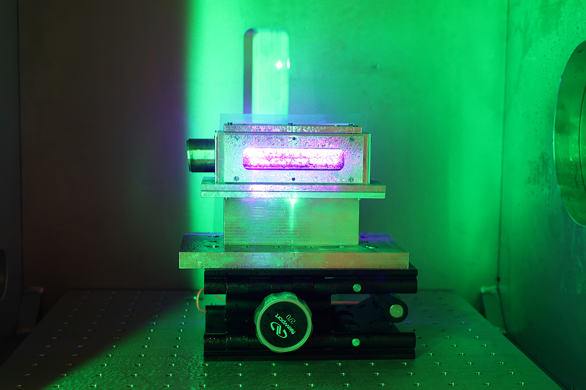 A piece of scientific equipment lit from the outside by green light. In a window in the side of the equipment, a pink light glows.