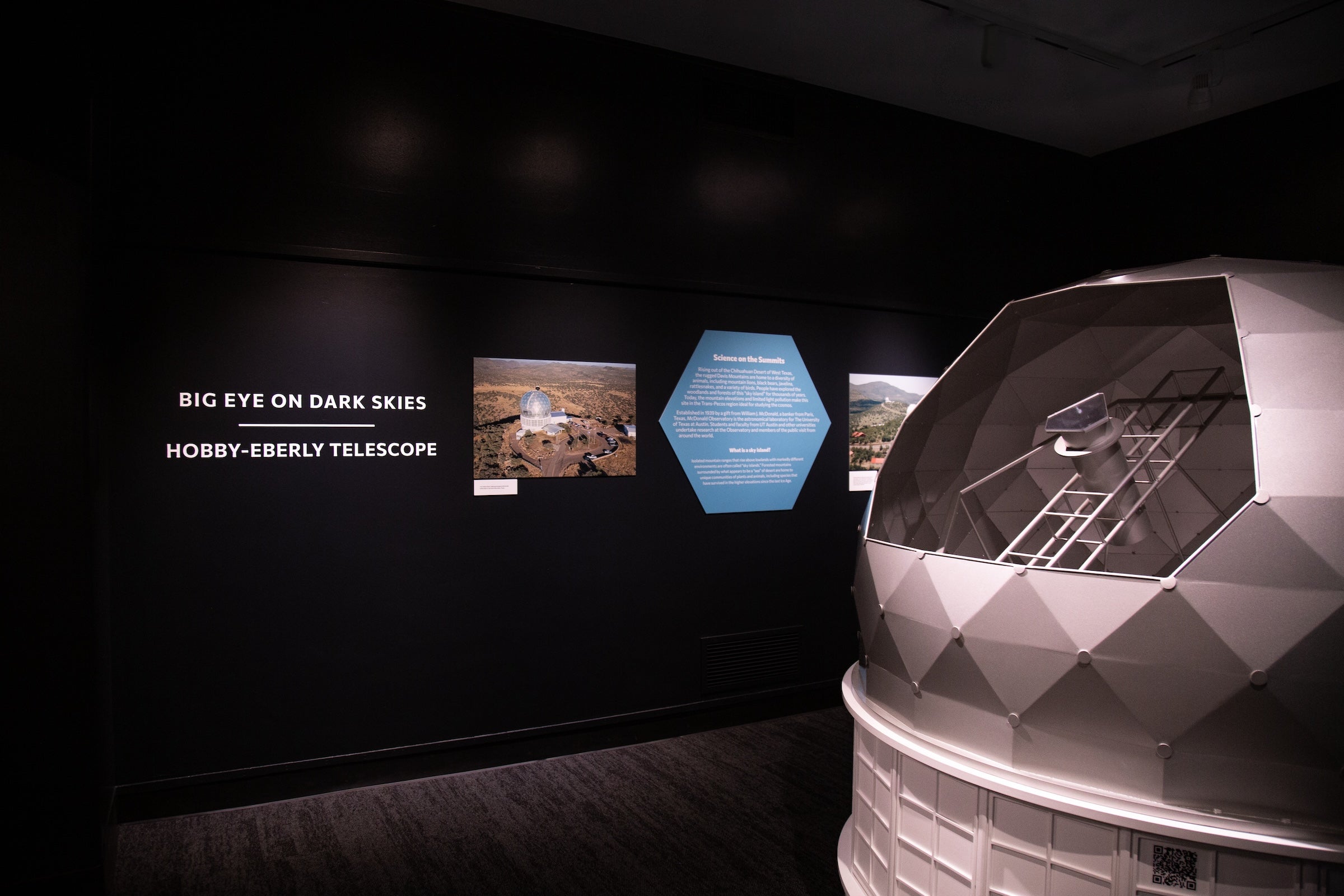 A dark room has museum-quality installations on its wall that read, "Big Eye on Dark Skies: Hobby-Eberly Telescope" near a 3D spherical telescope replica.