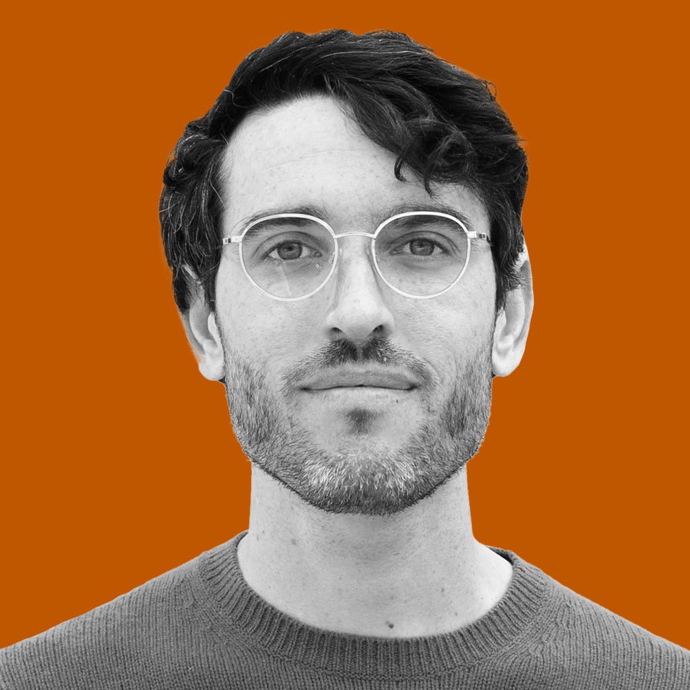 A young man with wireframe glasses and a light beard smiles while wearing a sweater.
