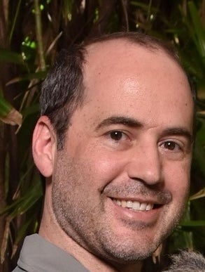 A man smiles for the camera in front of a background of leafy branches.