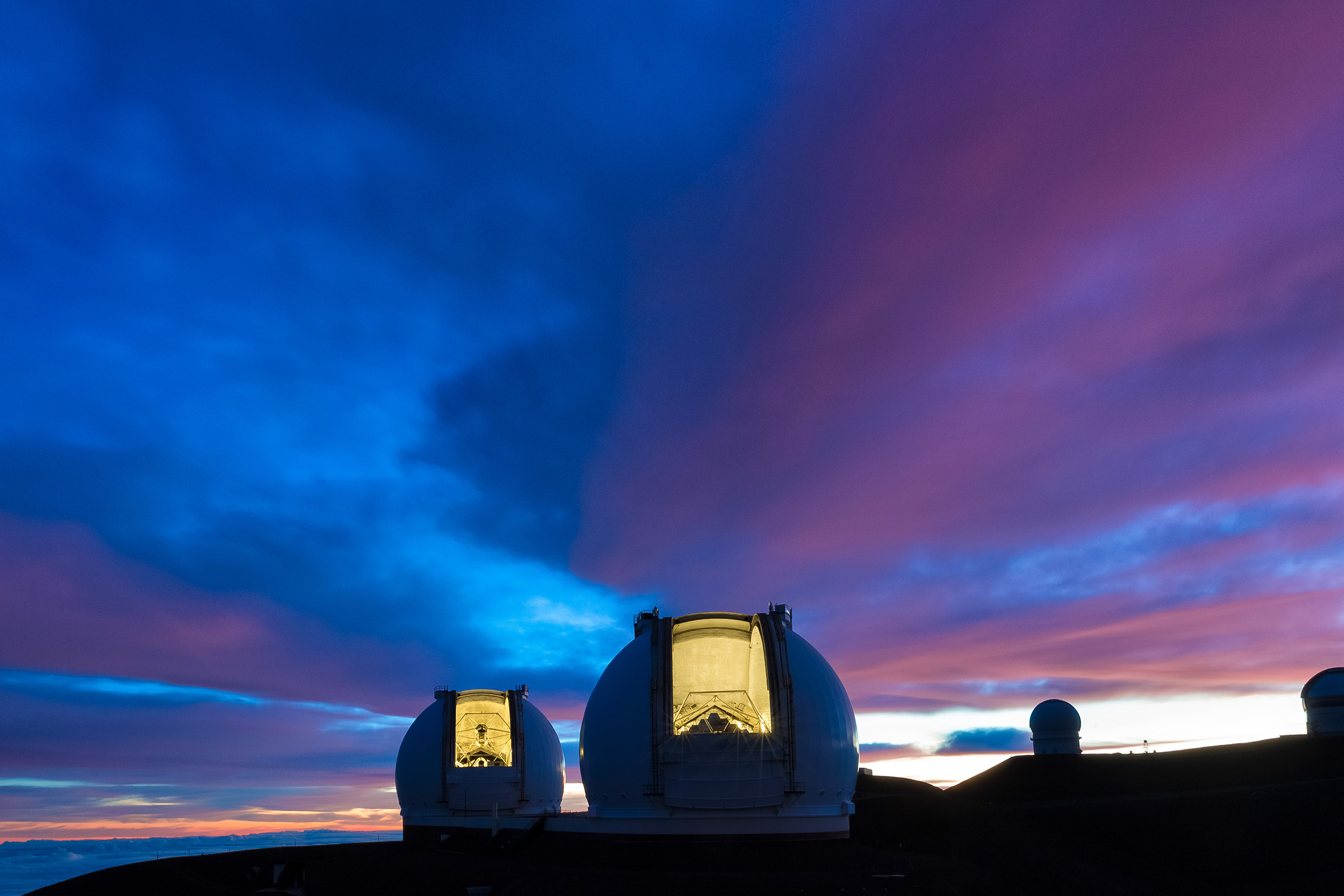 Two telescope domes silhouetted by sunlight creeping over the horizon beneath a blue and purple sky