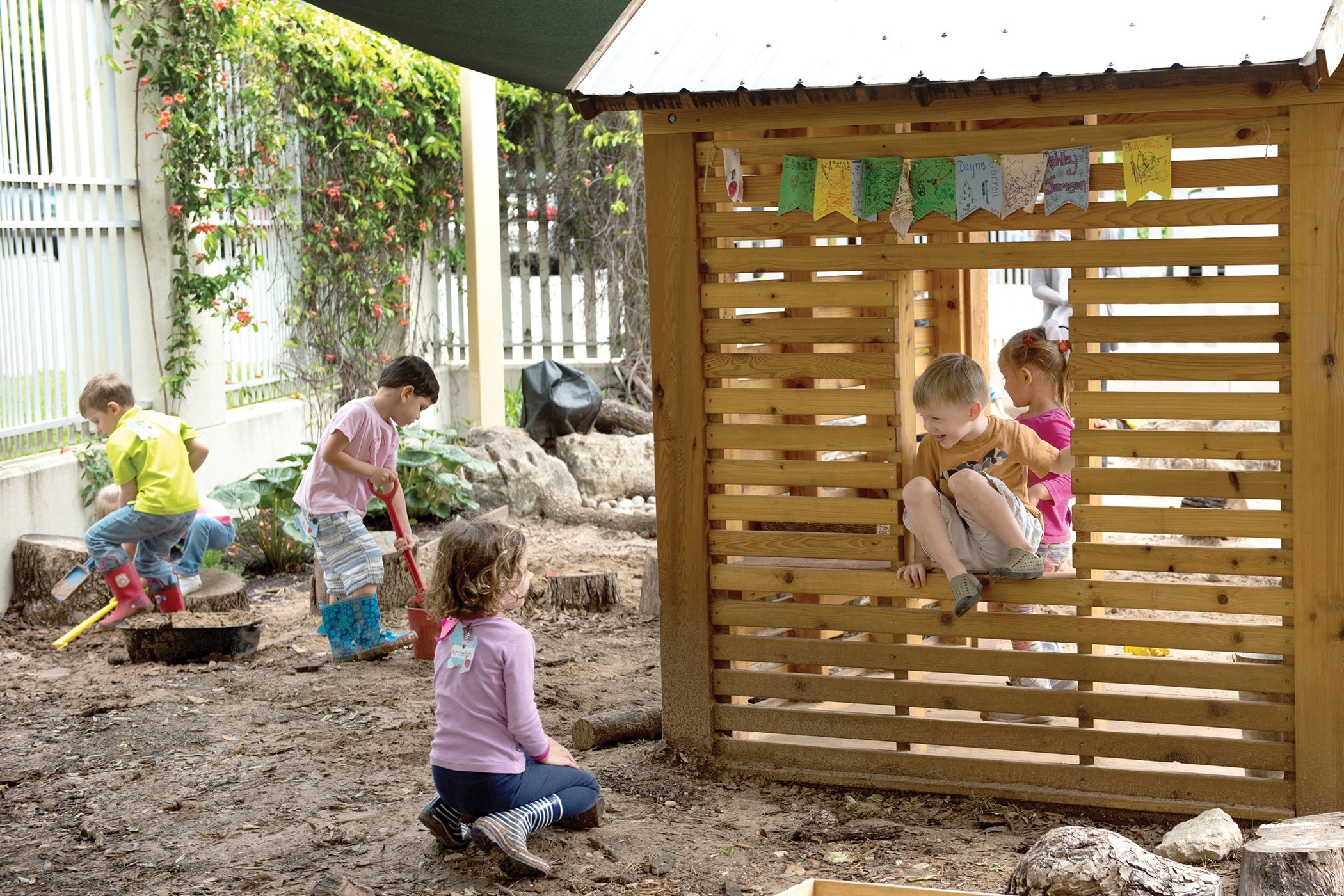 Five children at an outdoor preschool play with shovels and a house structure