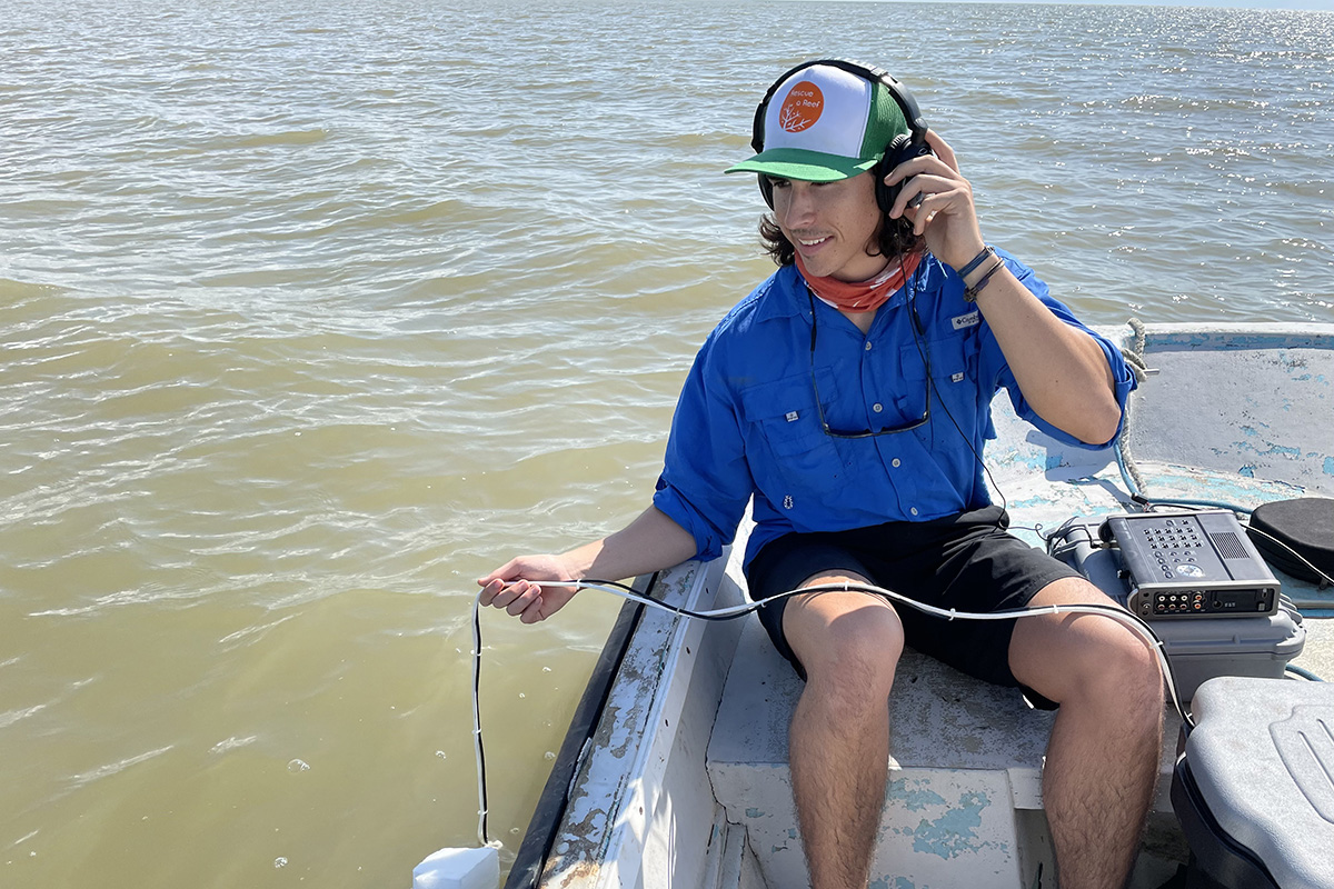 A scientist in a boat wearing headphones and dangles a microphone in the water