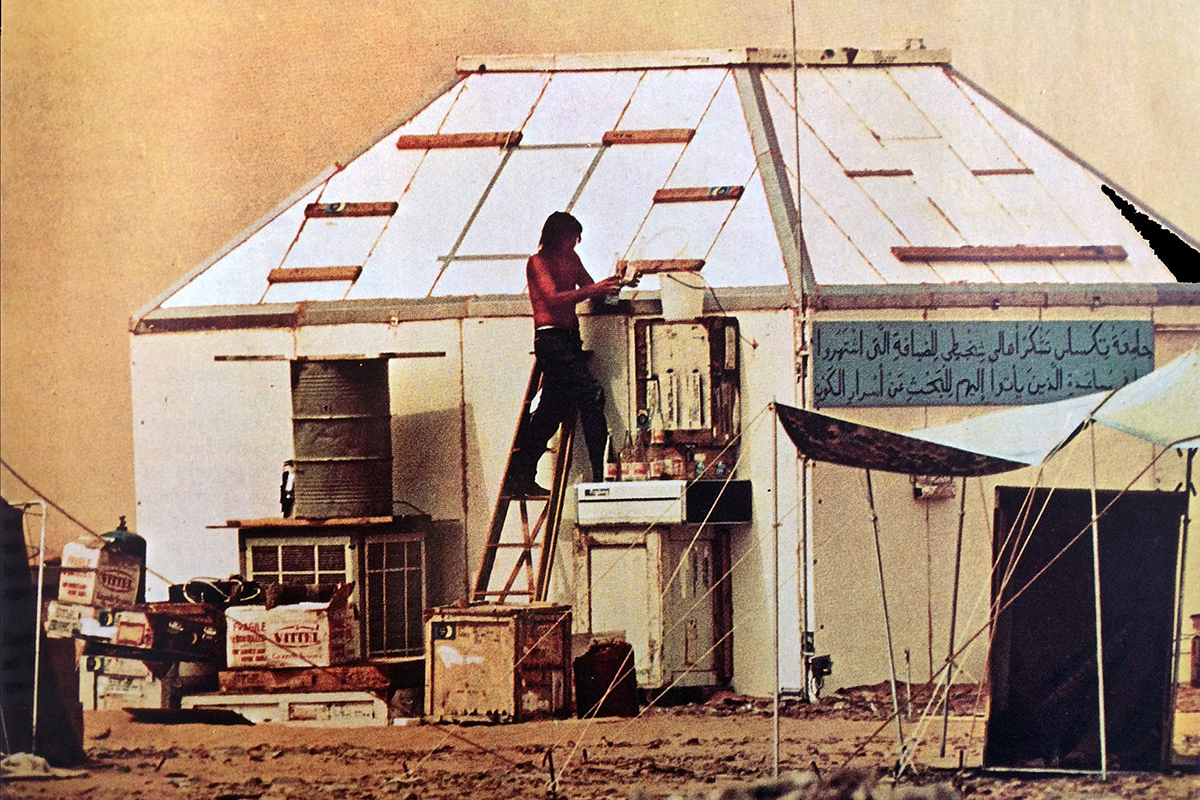 A man on a ladder works on a small white building in a desert