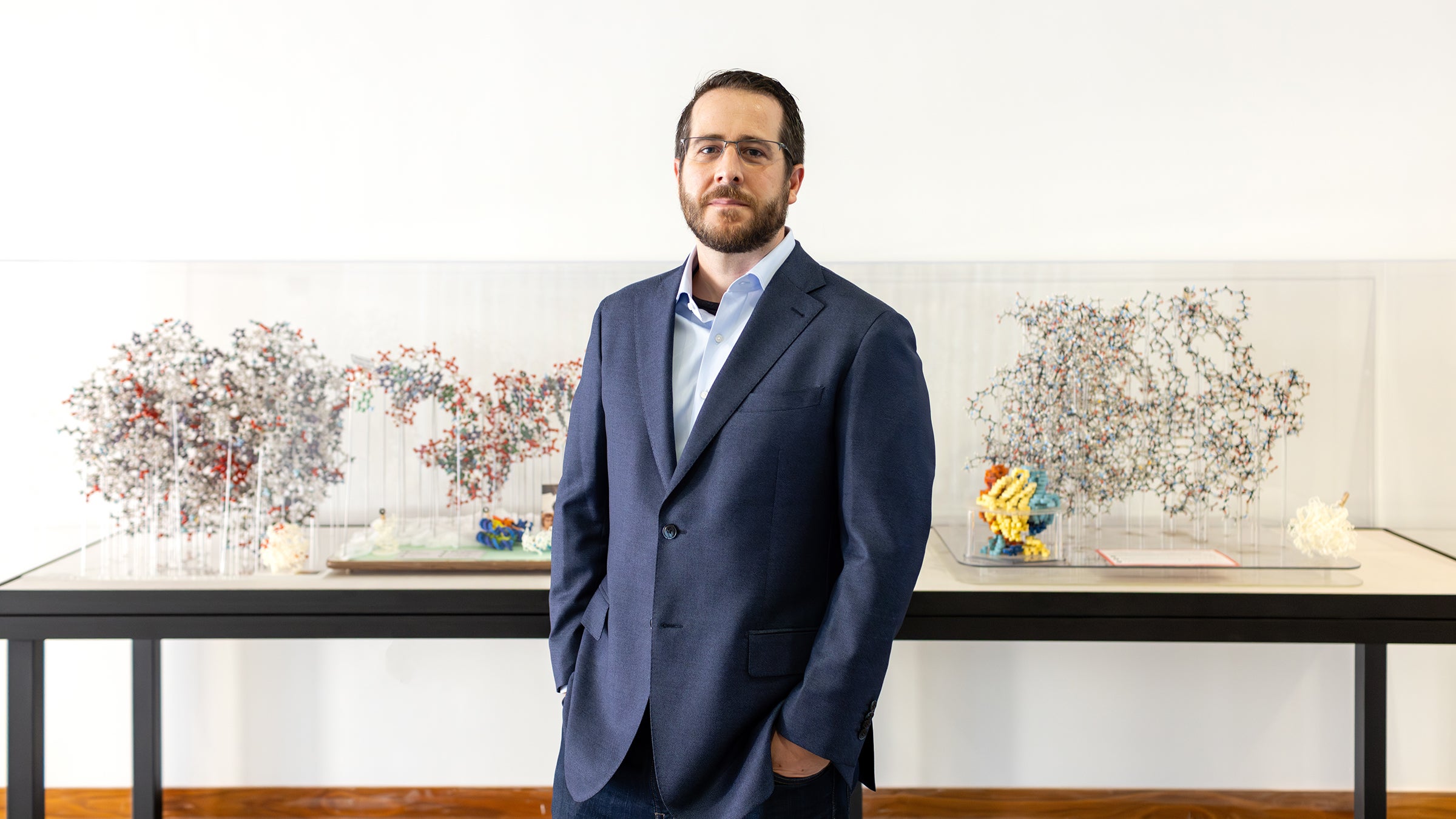 Portrait of Jason McLellan in a suit, standing in front of three-dimensional displays of proteins