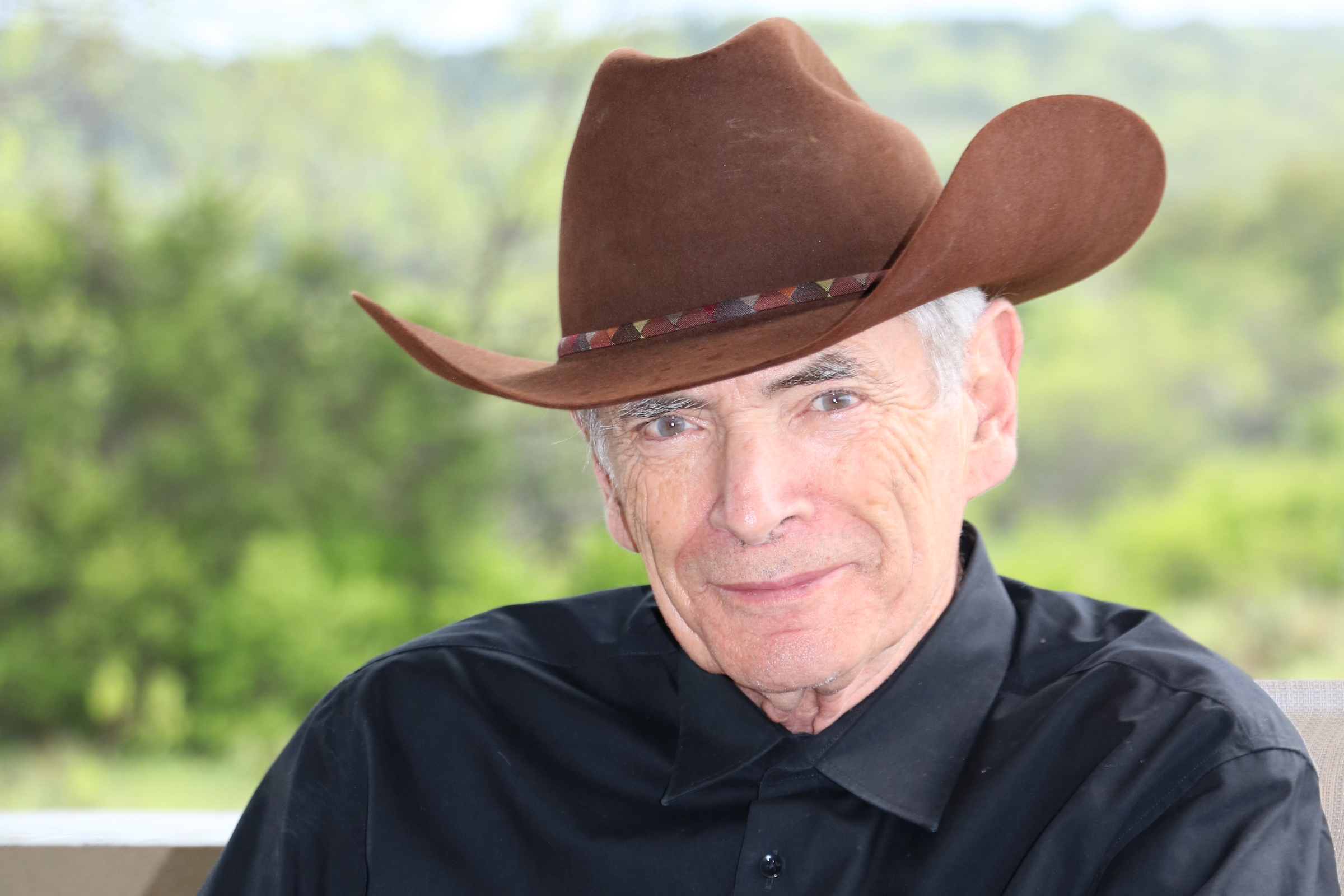 A man in a cowboy hat and collared black shirt smiles with trees and hills in the background