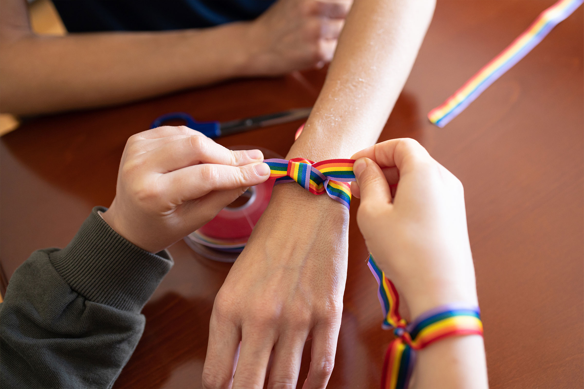 A pair of hands ties a rainbow ribbon on the wrist of another arm