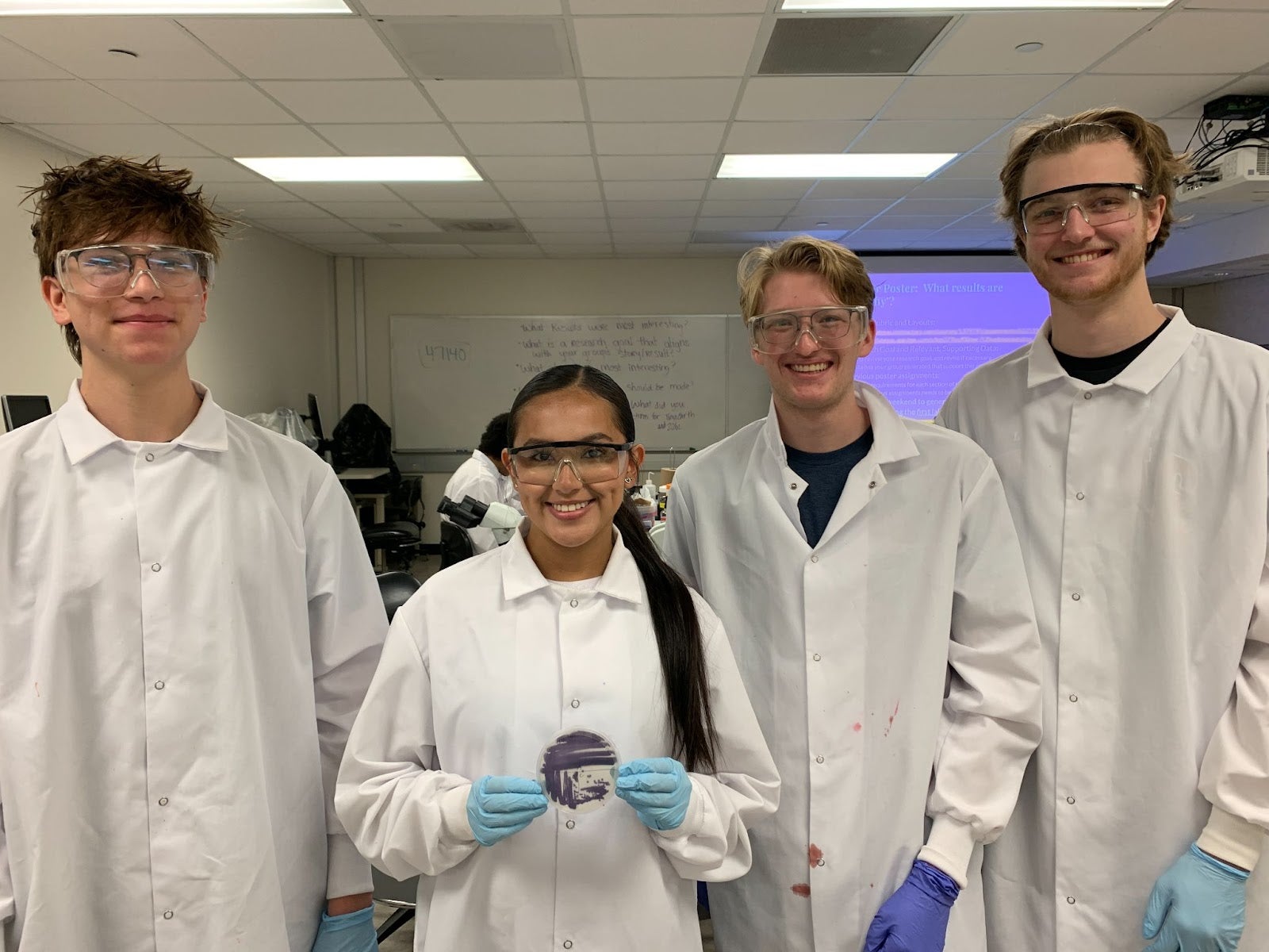 Four students wearing PPE smile for the camera in a classroom environment. A woman in the middle with a long ponytail holds up a petri dish.