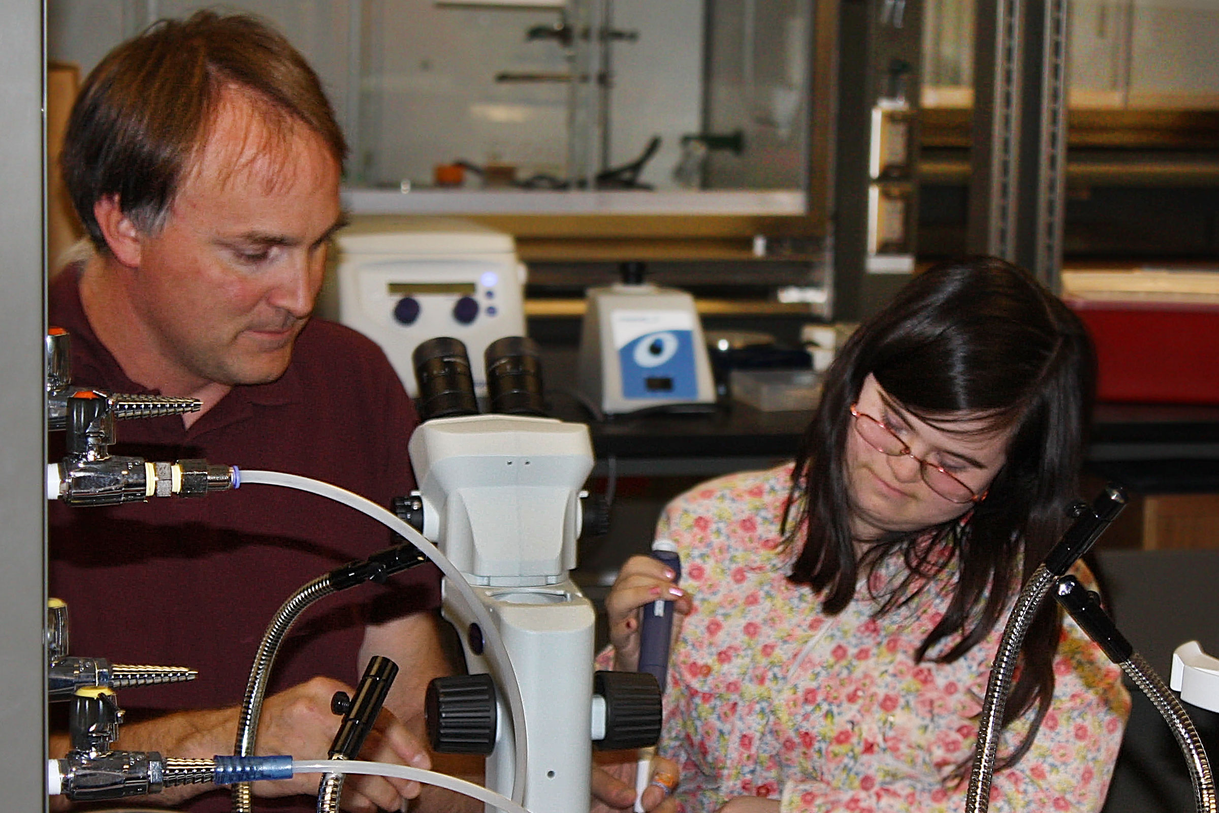 A scientist looks on in a lab as a student with developmental disabilities operates equipment.