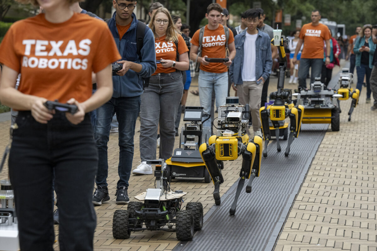 Several students and researchers in Texas Robotics t-shirts and holding controllers accompany a variety of robots walking and rolling down Speedway