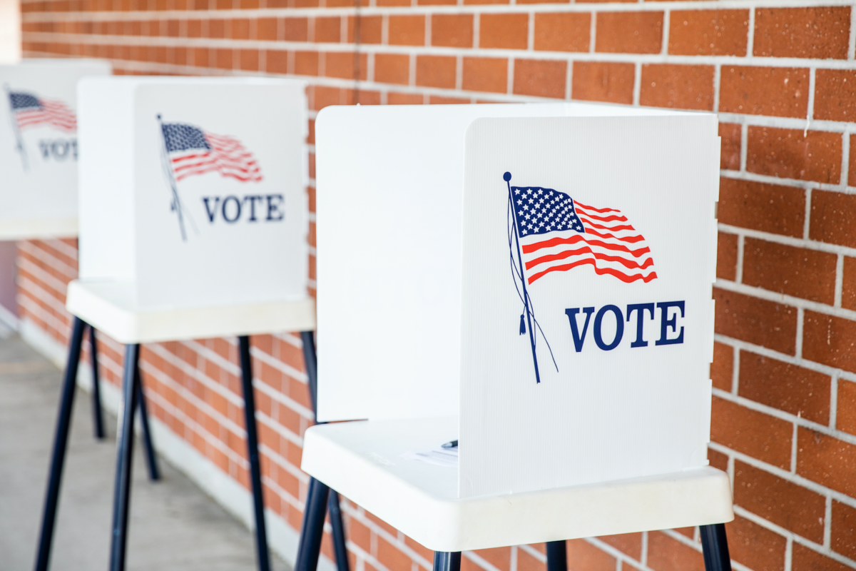 A row of voting booths sporting American flags and the word "vote" align against a brick wall.
